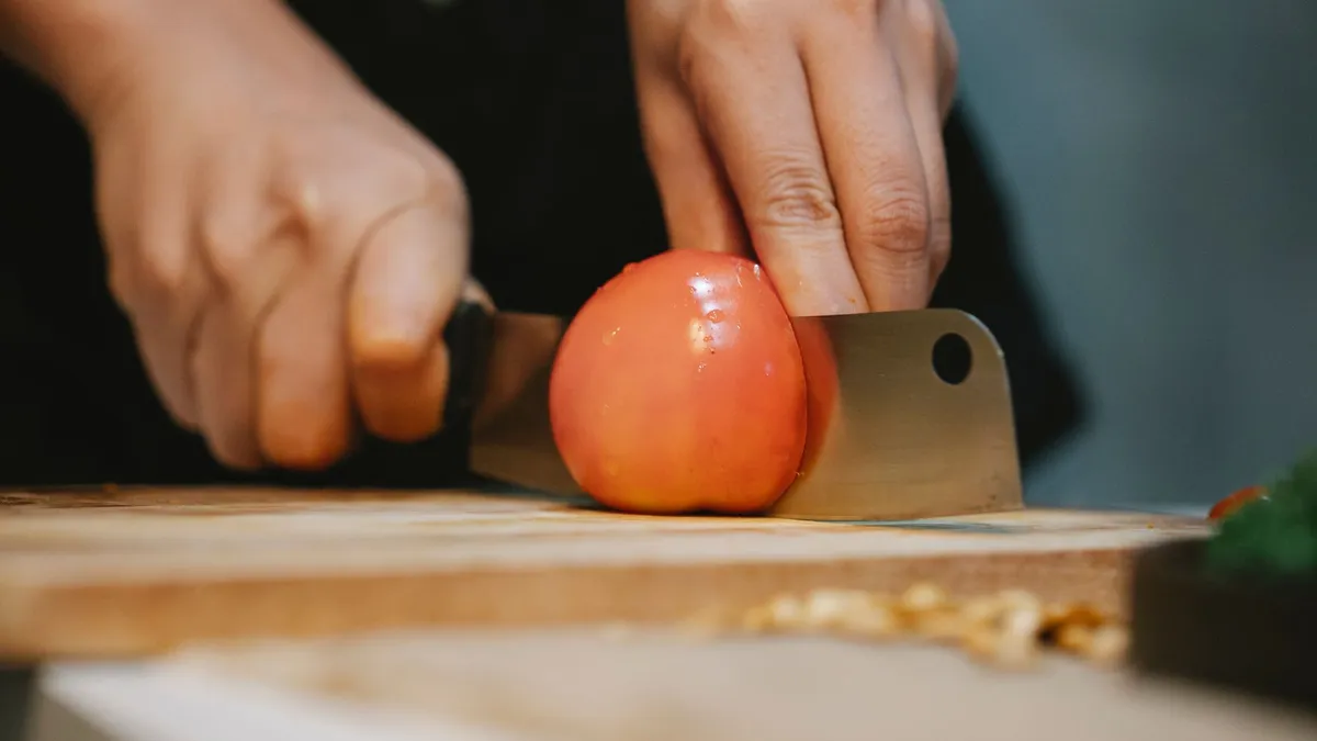 Crop unrecognizable chef slicing ripe wet red tomato with knife on wooden cutting board for cooking healthy dish