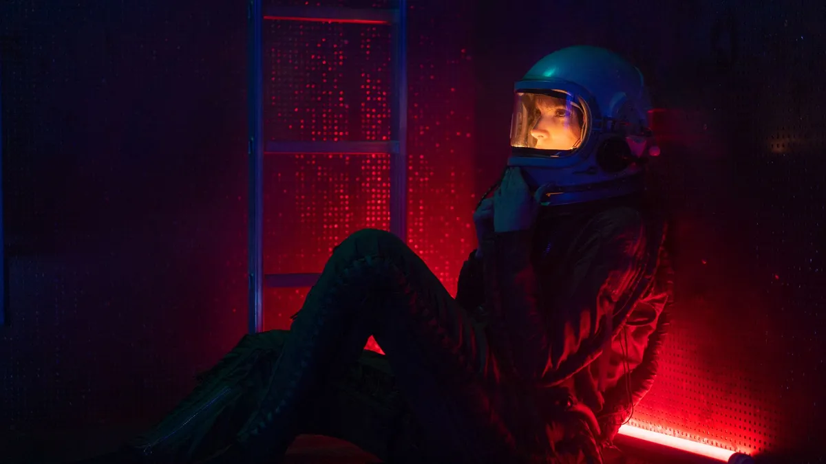 Woman astronaut in space suit sitting under red light, depicting future exploration. — Photo by Mikhail Nilov on Pexels